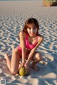 A woman in a pink bikini sitting on a beach with a coconut.